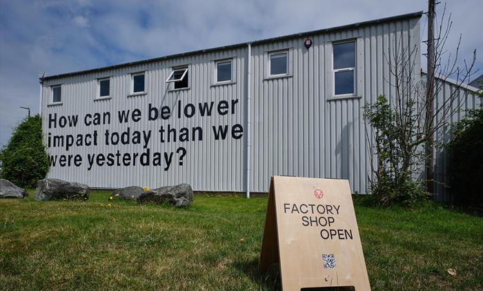 The exterior of the Hiut Denim factory features bold black text reading “How can we be lower impact today than we were yesterday?” A wooden sign on th